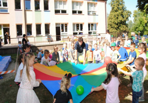 w ogrodzie dzieci trzymają chustę animacyjną i poruszają nią tak aby umieszczone na niej balony zmieniały miejsce